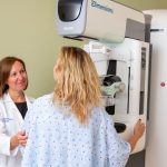 Dr. Alison Chetlen, left, touches a patient’s arm as she positions her in front of a mammography machine. Chetlen is wearing a white coat with her name and “Radiology” on the upper-right.