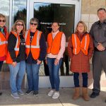 Six people - five wearing high-visibility vests, one wearing a police uniform - stand in front of the building.