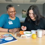 Two women sit at a table looking at a plate of food. A diabetes booklet is on the left. The woman on the left is wearing a knit top, glasses and a name badge. The woman on the right is wearing a long-sleeved top and is holding a cherry tomato.